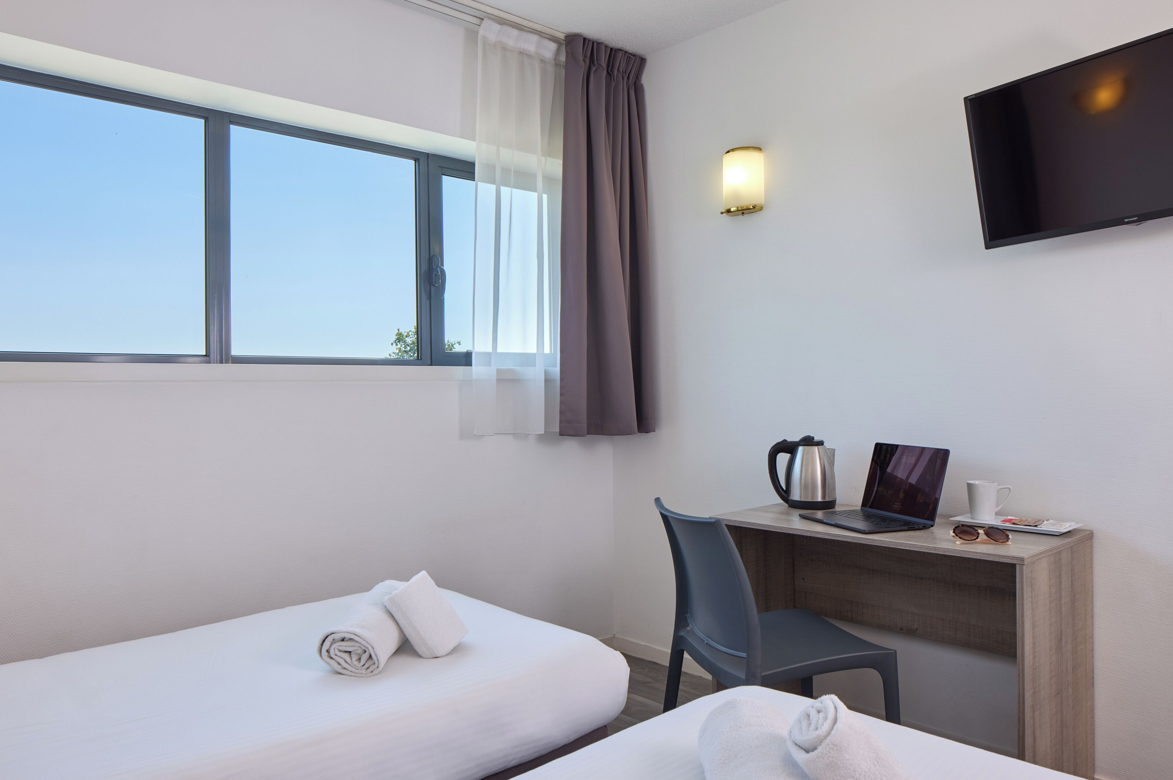 Habitación de hotel con dos camas individuales, un escritorio con una computadora portátil, un cafetera y un televisor instalado en la pared.
