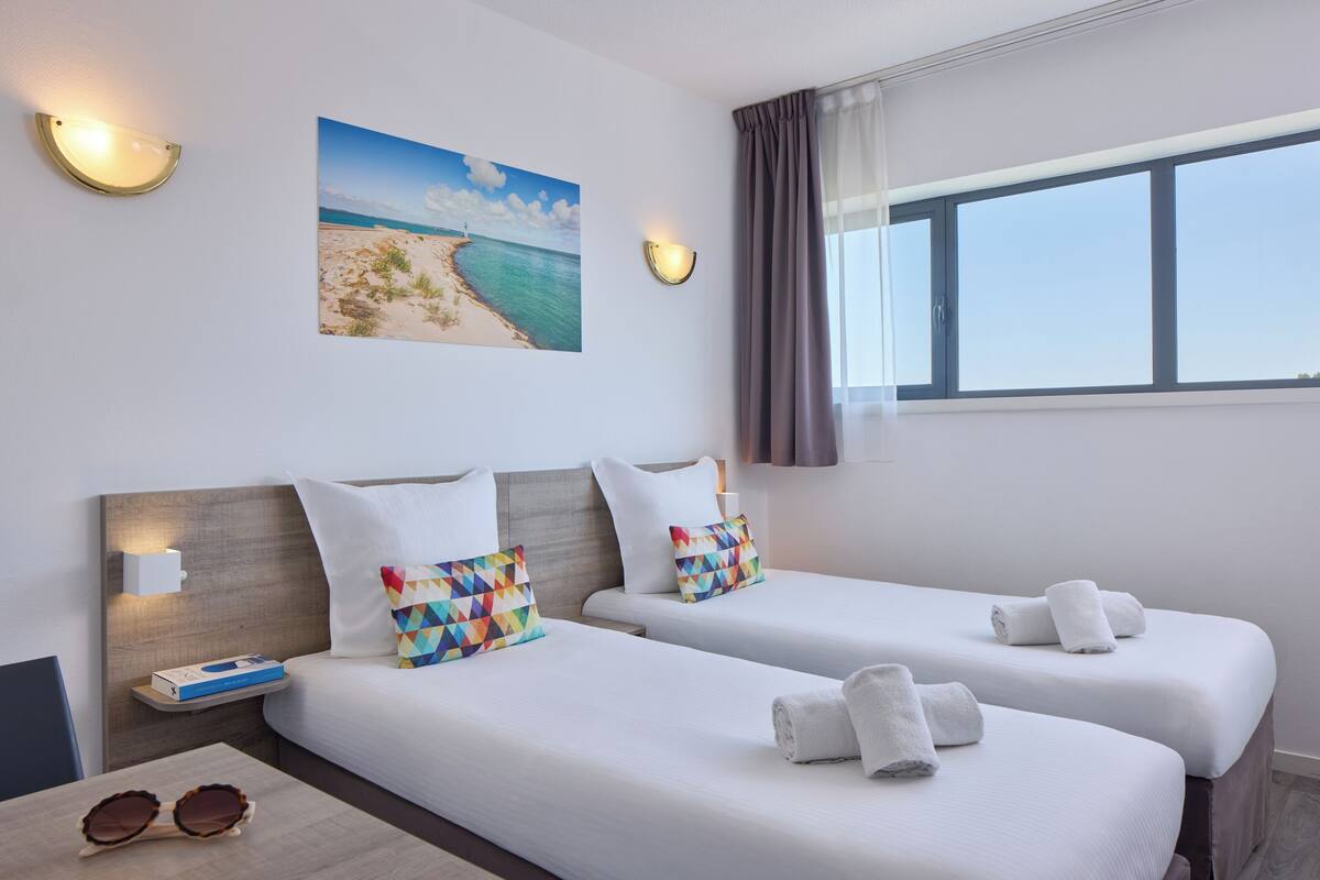Una habitación de hotel con dos camas, un cuadro de una playa, una ventana con cortinas y una mesita de noche con un libro y gafas.