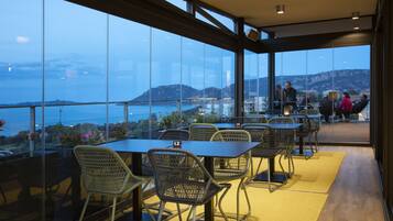 Un restaurante con amplios ventanales que ofrece vistas al mar y a las montañas. Mesas y sillas dispuestas para comer.
