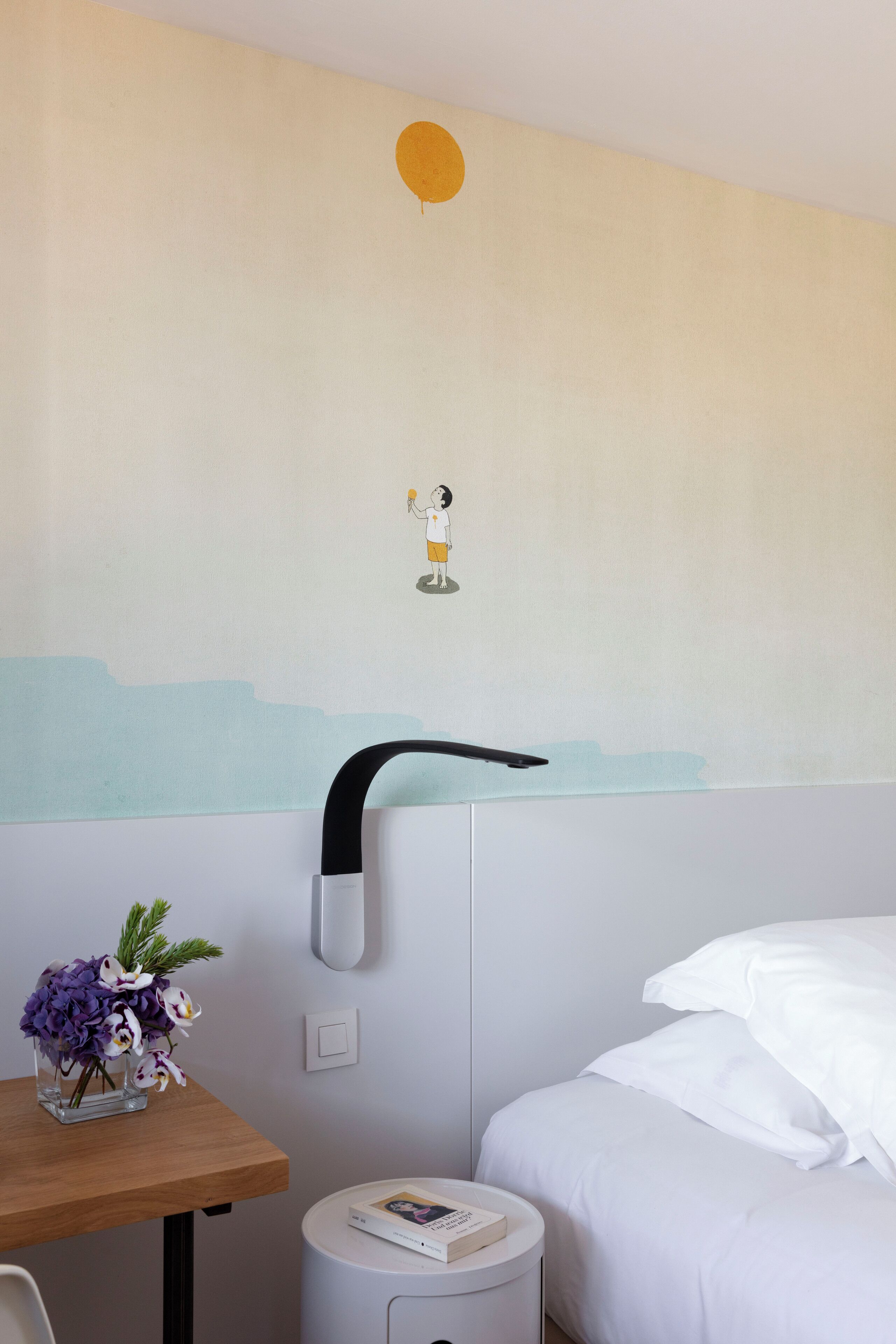 Une chambre d’hôtel avec un lit, une petite table sur laquelle est posé un vase de fleurs, une table de chevet ronde et blanche, et un mur orné d’une simple fresque.