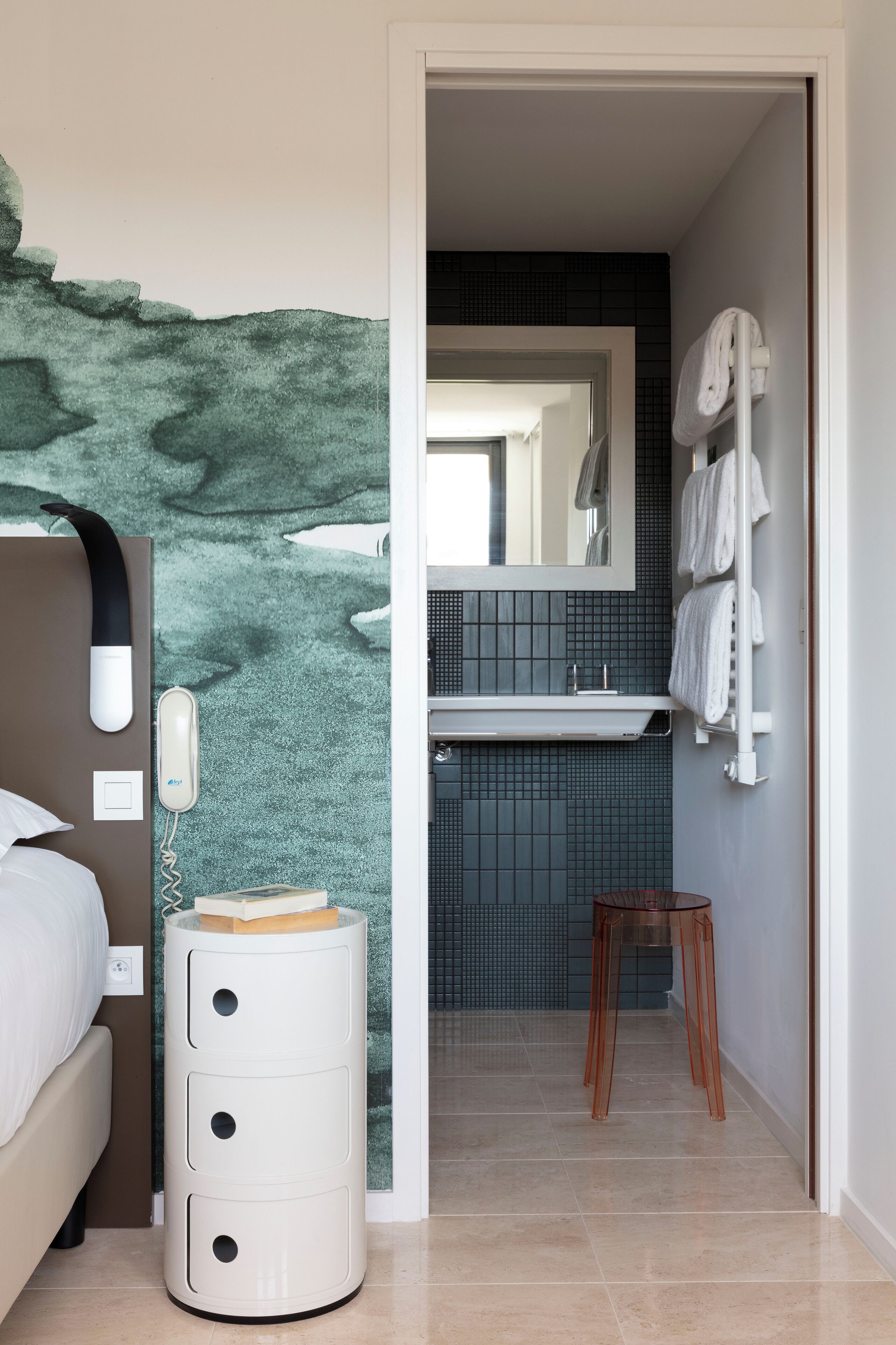 A hotel room with a bed, a bedside phone, a small table with a book, a mirror, a sink, and a towel rack.