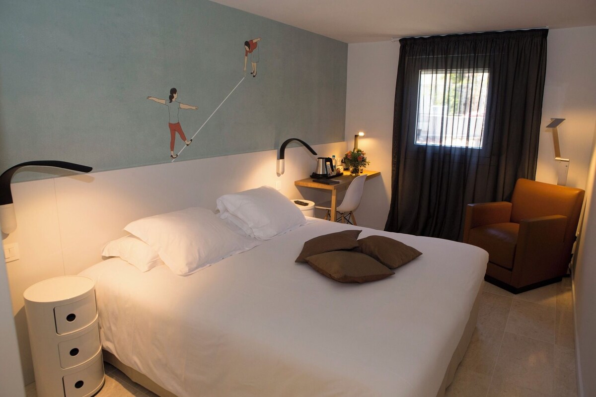 Ein Hotelzimmer mit Bett, Schreibtisch, Stuhl, Lampe und Wandbild.