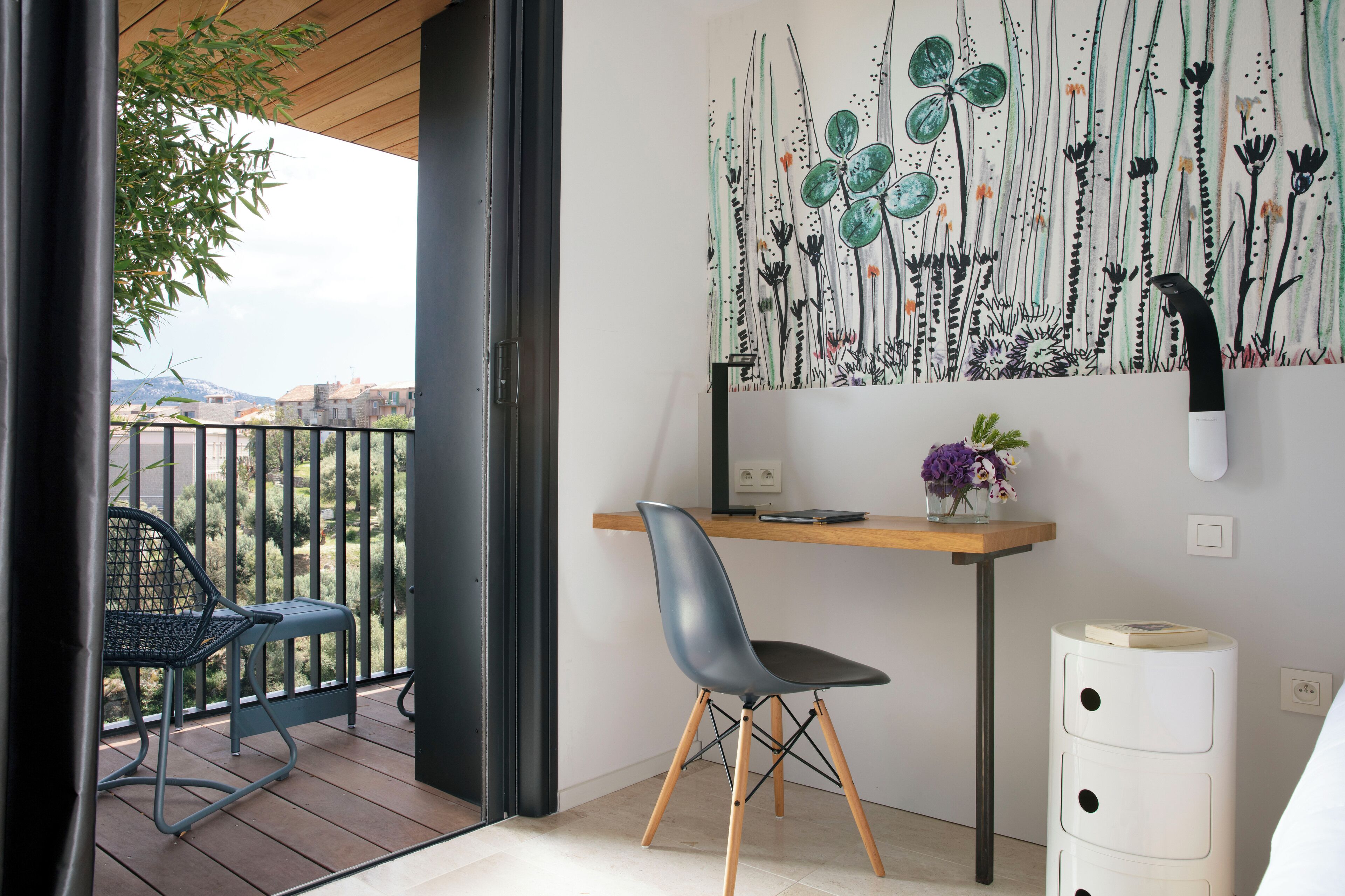 A balcony with a chair, a desk with a chair, a wall with a botanical painting, and a white cabinet.