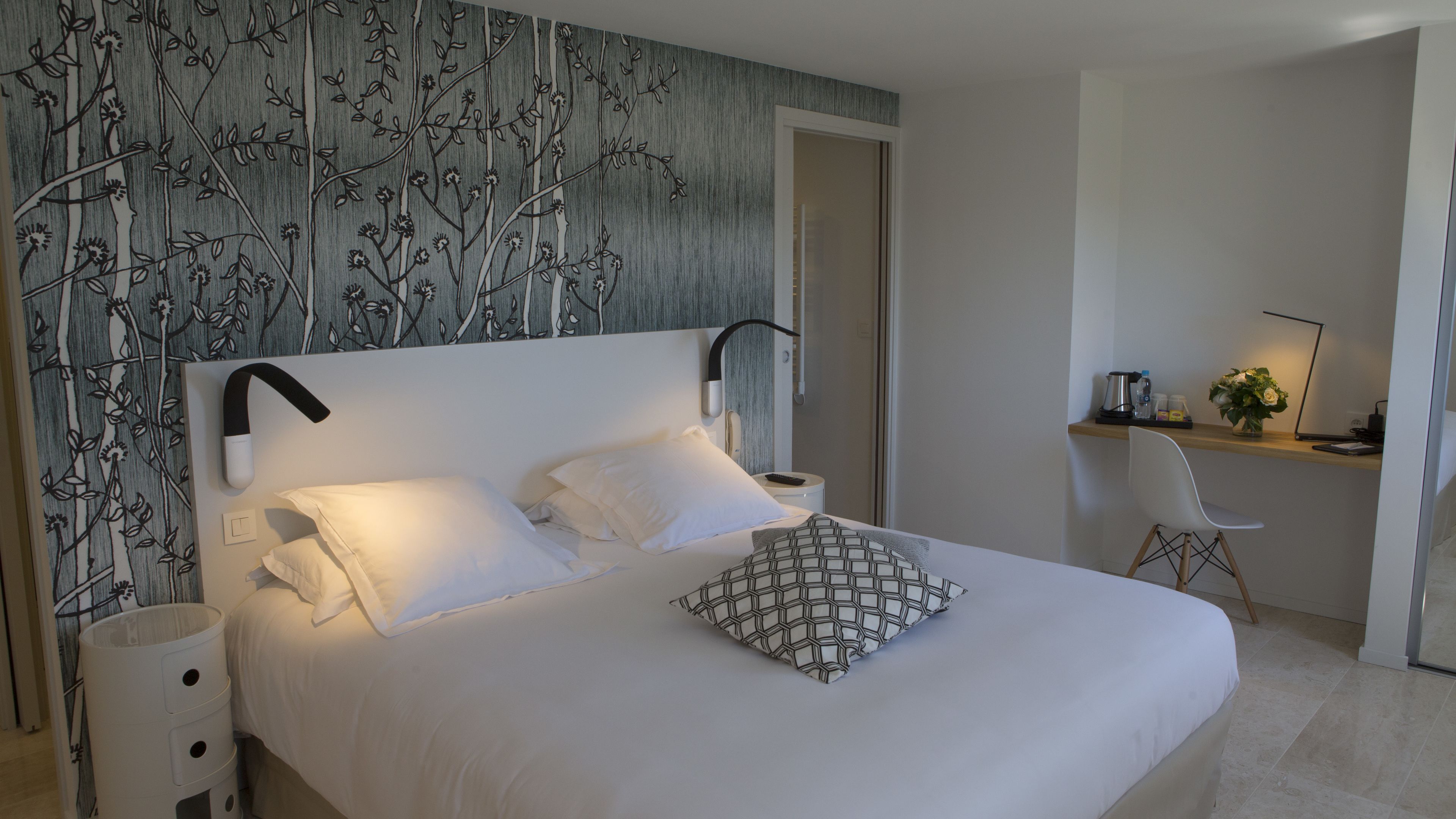 A modern bedroom with a large bed, white bedding, a patterned pillow, a bedside table with a lamp, and a desk with a chair.