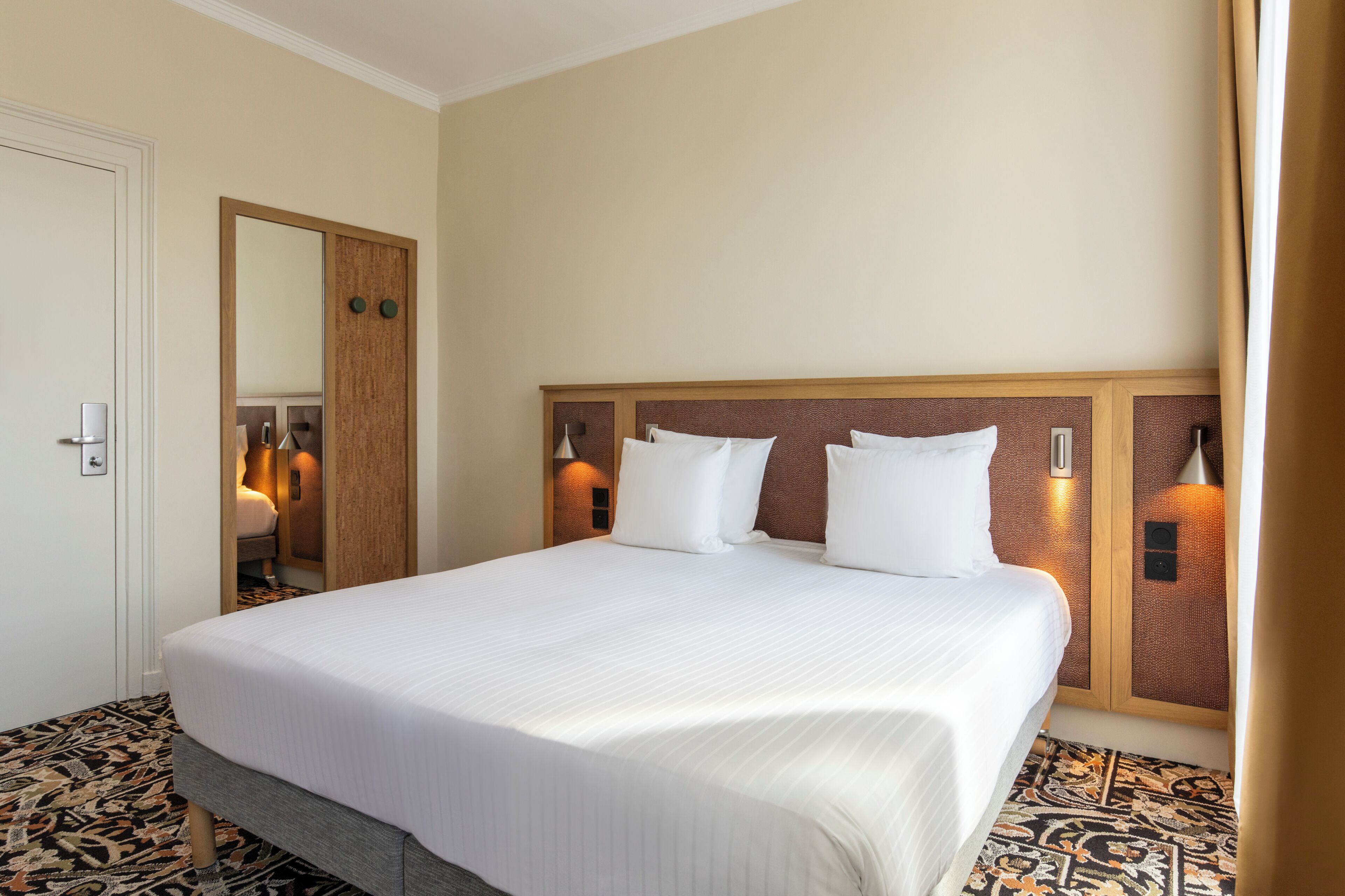 A hotel room with a large bed, a wooden headboard, two wall-mounted lamps, and a patterned carpet.