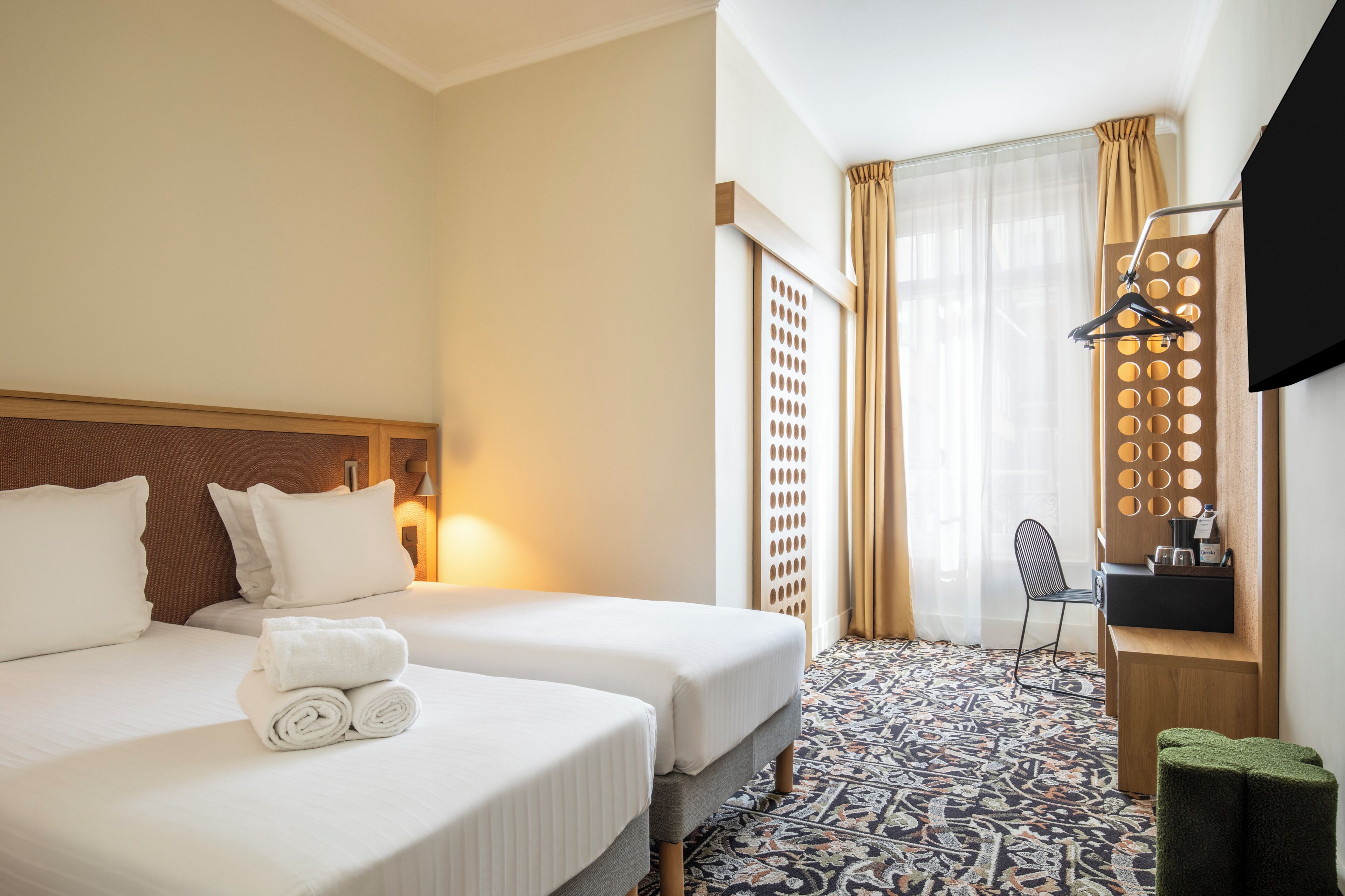 A hotel room with two beds, a patterned floor, a desk with a chair, and a wall-mounted TV.