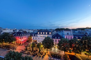 A cityscape at dusk with illuminated buildings and a central square with outdoor seating.
