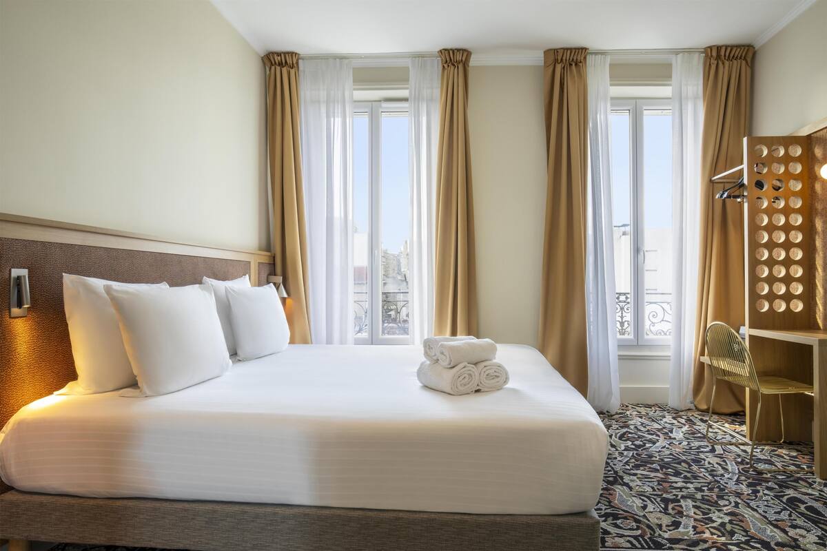 A hotel room with a large bed, a wooden headboard, a patterned carpet, and a view of the city through the windows.