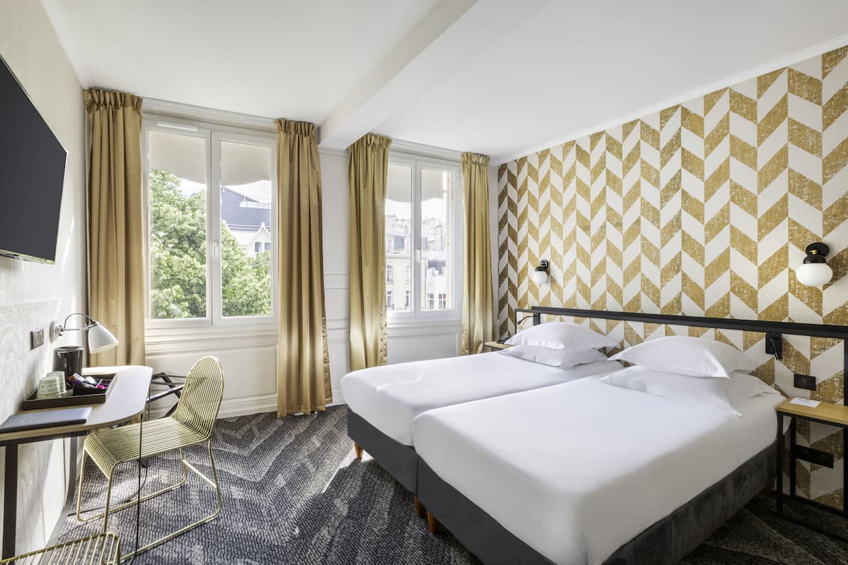 Habitación de hotel con dos camas, un escritorio con silla, televisor y ventanas amplias con cortinas.