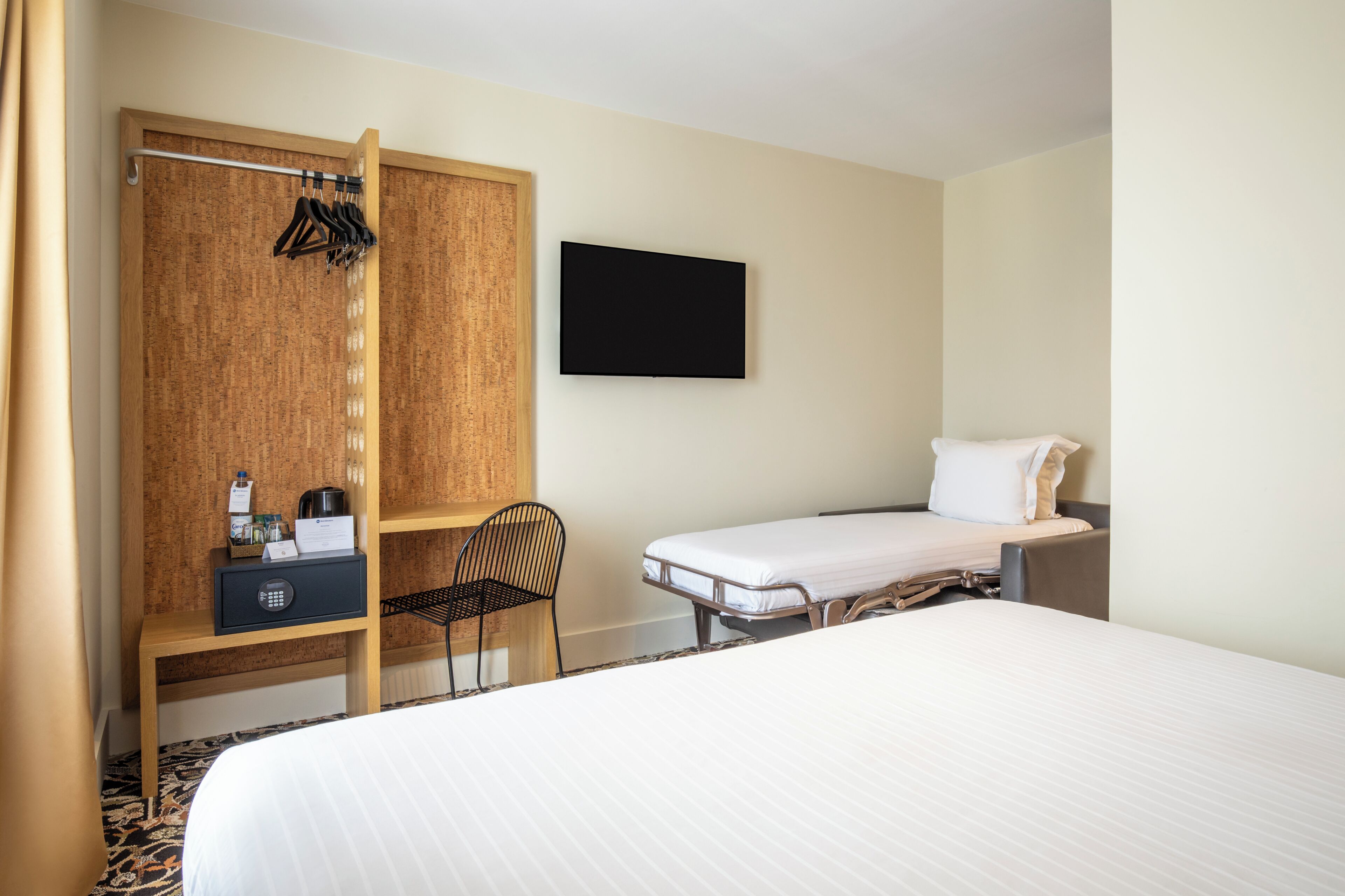 A hotel room with a bed, a desk with a chair, a television mounted on the wall, and a wardrobe with a mirror.