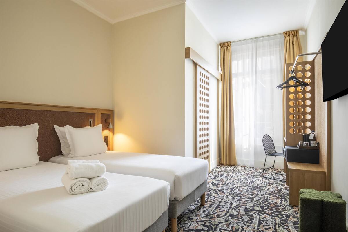 A hotel room with two beds, a patterned floor, a desk with a mirror, and a hanging coat rack.