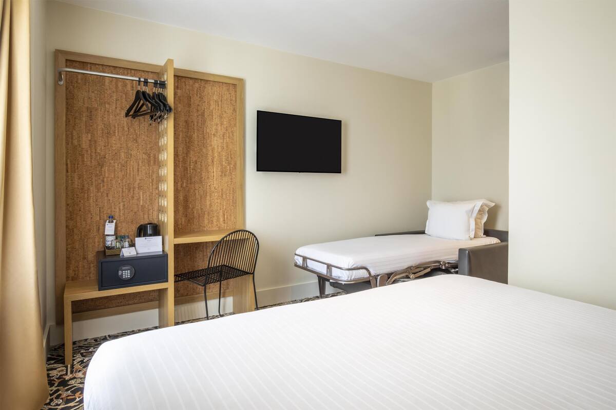 A hotel room with a bed, a desk with a chair, a wardrobe, a television, and a small bedside table.