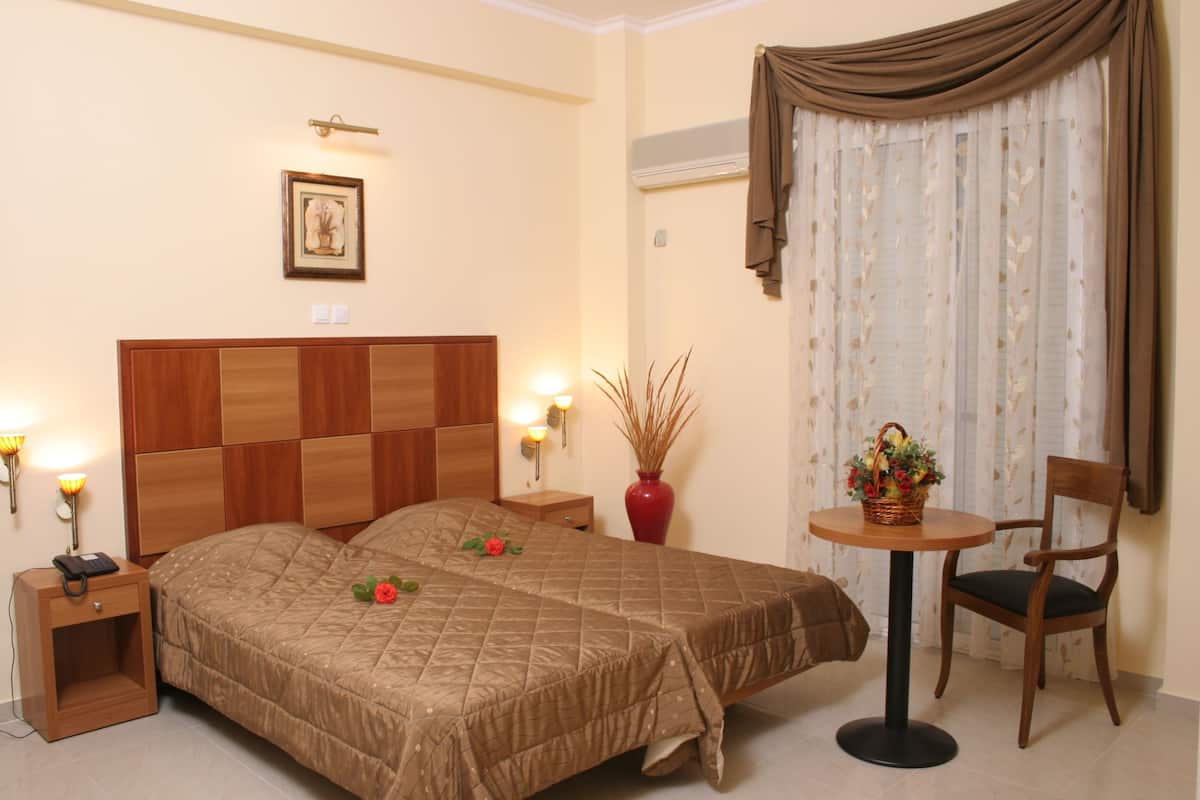 A hotel room with a wooden headboard, a bed with a brown quilt, a nightstand with a lamp, a red vase, and a small round table with a chair.