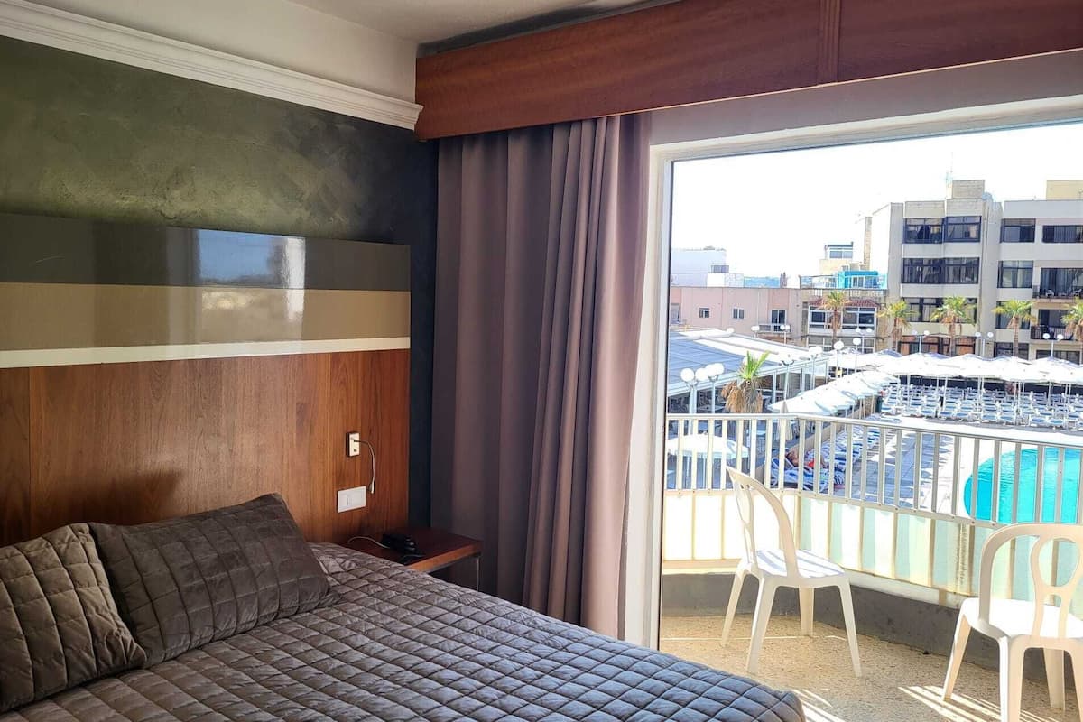 Habitación de hotel con cama, mesita de noche y ventana que ofrece vistas a una piscina y edificios.