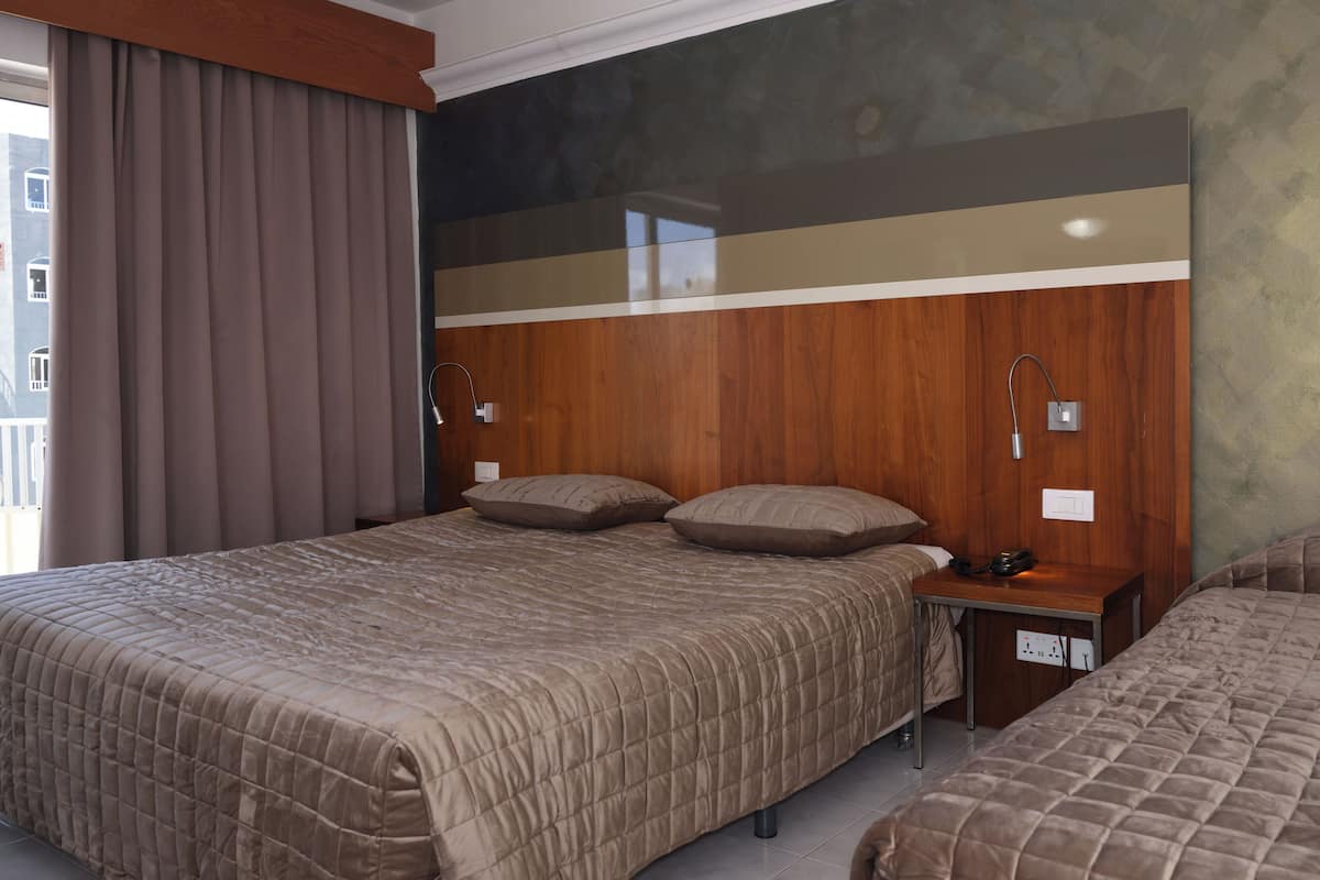 Habitación de hotel con dos camas, cabeceros de madera, una mesita de noche con lámpara y vistas al exterior.