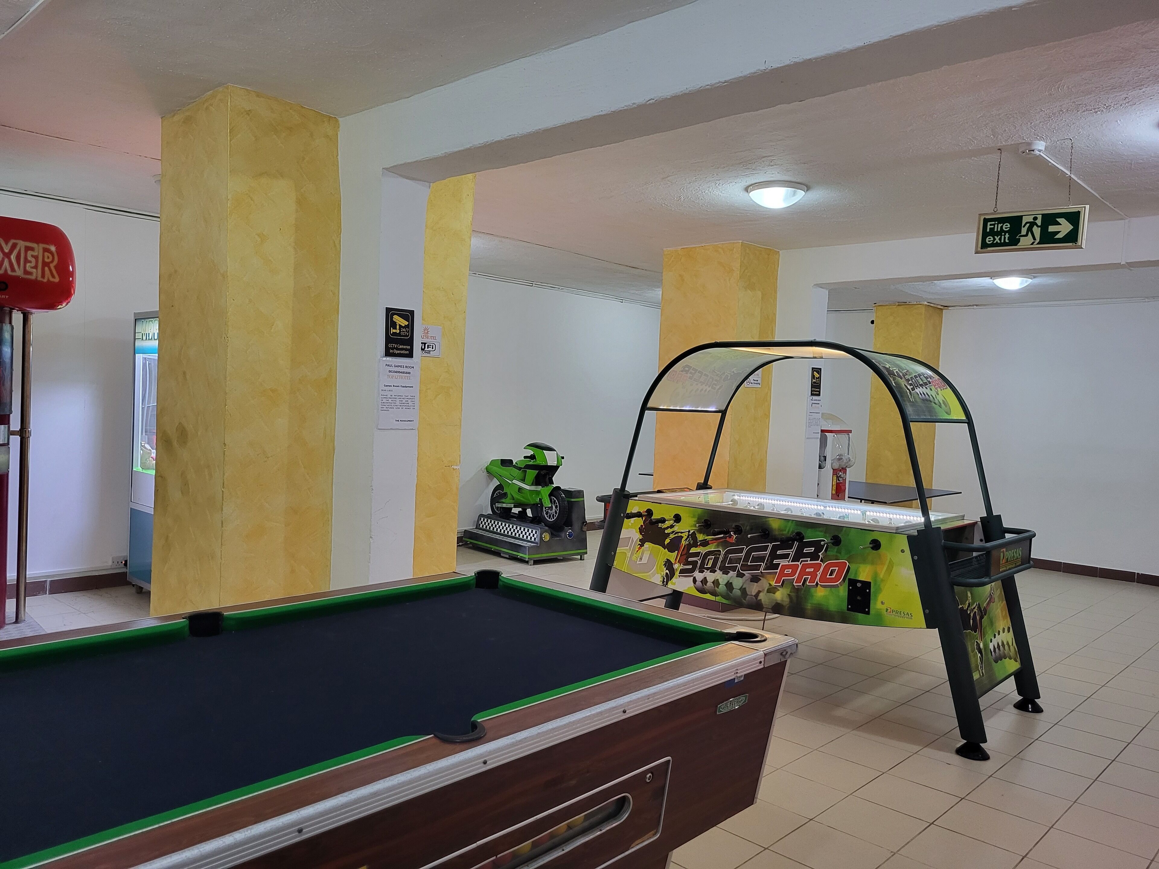A pool table in a room with yellow pillars, a fire exit sign, and a green arcade machine.