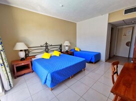 Habitación de hotel con dos camas, cada una con una colcha azul y almohadas amarillas. Hay dos mesitas de noche con lámparas, una silla de madera y una mesa de madera.