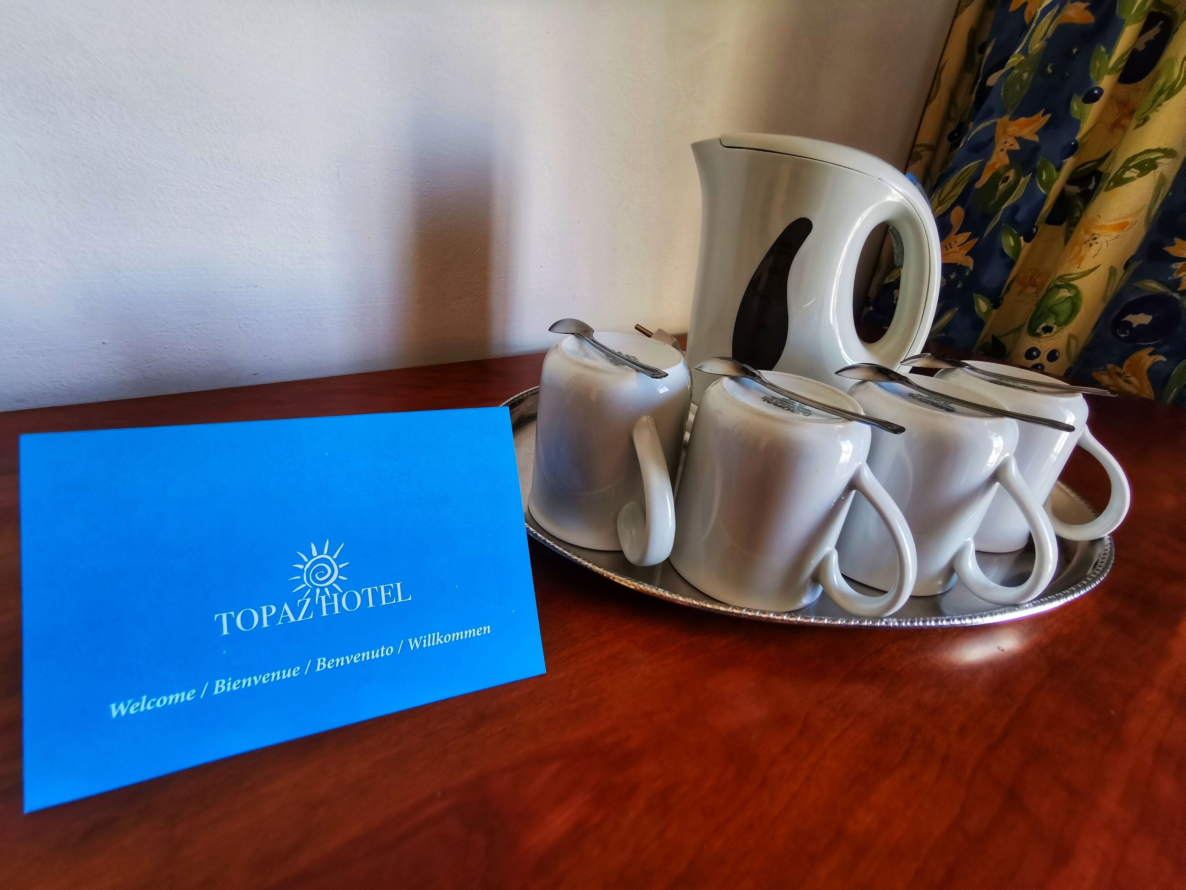 Un juego de tazas de cerámica blanca y una tetera sobre una bandeja, acompañadas de una tarjeta azul de bienvenida para Topaz Hotel.