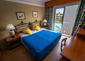 Habitación de hotel con cama, dos lámparas de noche, una mesita, un escritorio de madera y un balcón con vistas a la piscina.