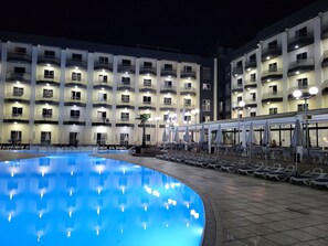 A swimming pool with lounge chairs and umbrellas, surrounded by a building with multiple balconies and windows. - Topaz Hotel (St. Paul's Bay)