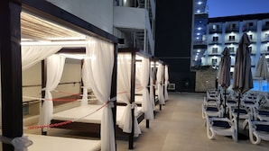 Una fila de camas individuales con cortinas blancas y cadenas rojas, un área de piscina con sillones de descanso y un edificio con balcones al fondo.