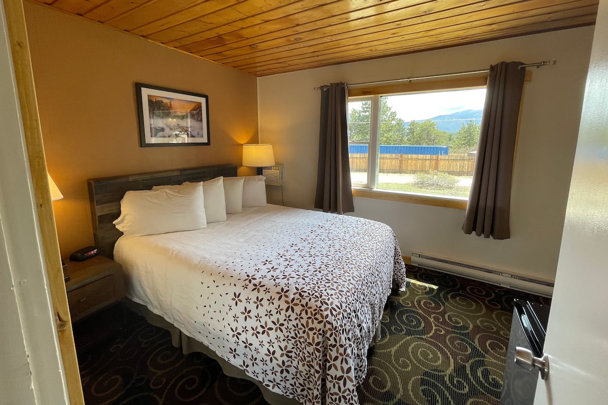 A hotel room with a wooden ceiling, a bed with a patterned bedspread, a nightstand with a lamp, a window with curtains, and a view of the outdoors.