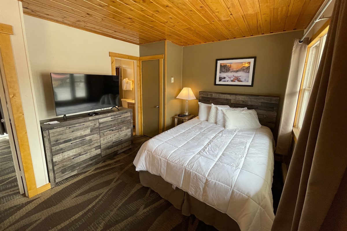 A hotel room with a wooden ceiling, a bed with white bedding, a TV on a wooden dresser, and a painting on the wall.