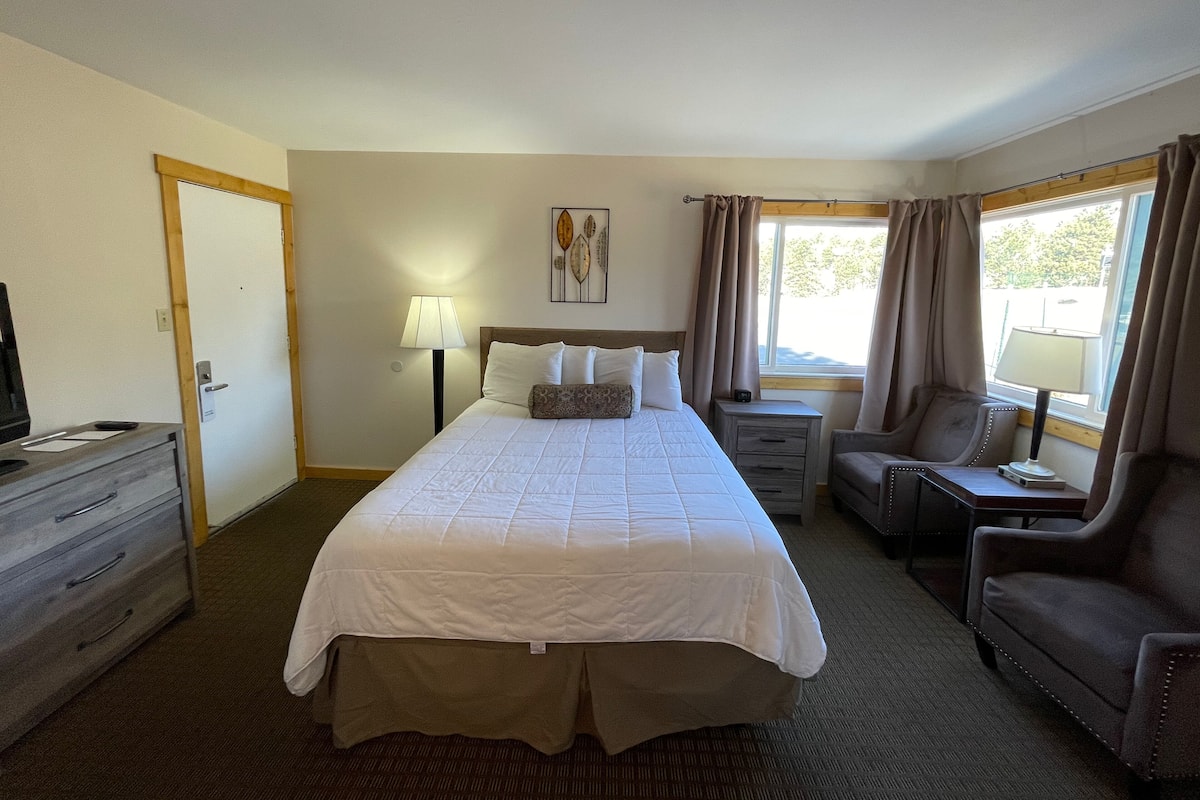 A hotel room with a large bed, a desk, a chair, a dresser, and a window with curtains.