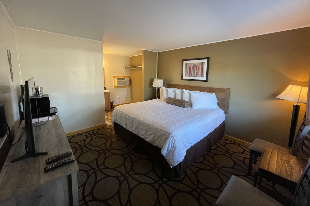A hotel room with a bed, a desk with a computer and remote controls, a chair, and a lamp.