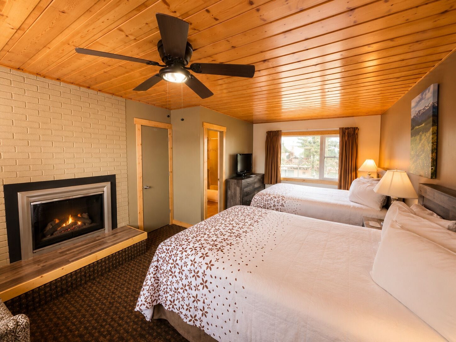 A hotel room with a fireplace, two beds, a ceiling fan, and a window with curtains.