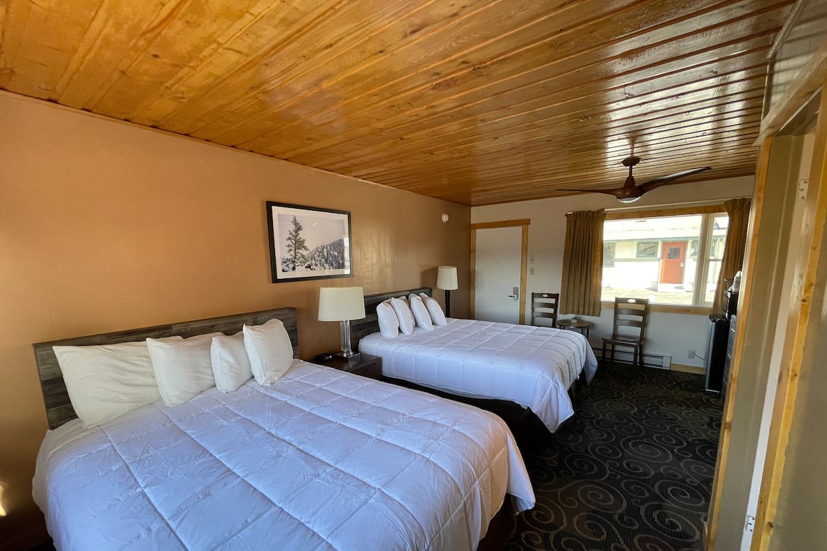 A hotel room with two beds, a wooden ceiling, a framed picture on the wall, a desk with a chair, and a window with curtains.