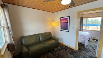 Ein Zimmer mit einem grünen Ledersofa, einem Deckenventilator, einem gerahmten Bild, einer Lampe und einem Fenster mit Blick auf eine Holzterrasse.