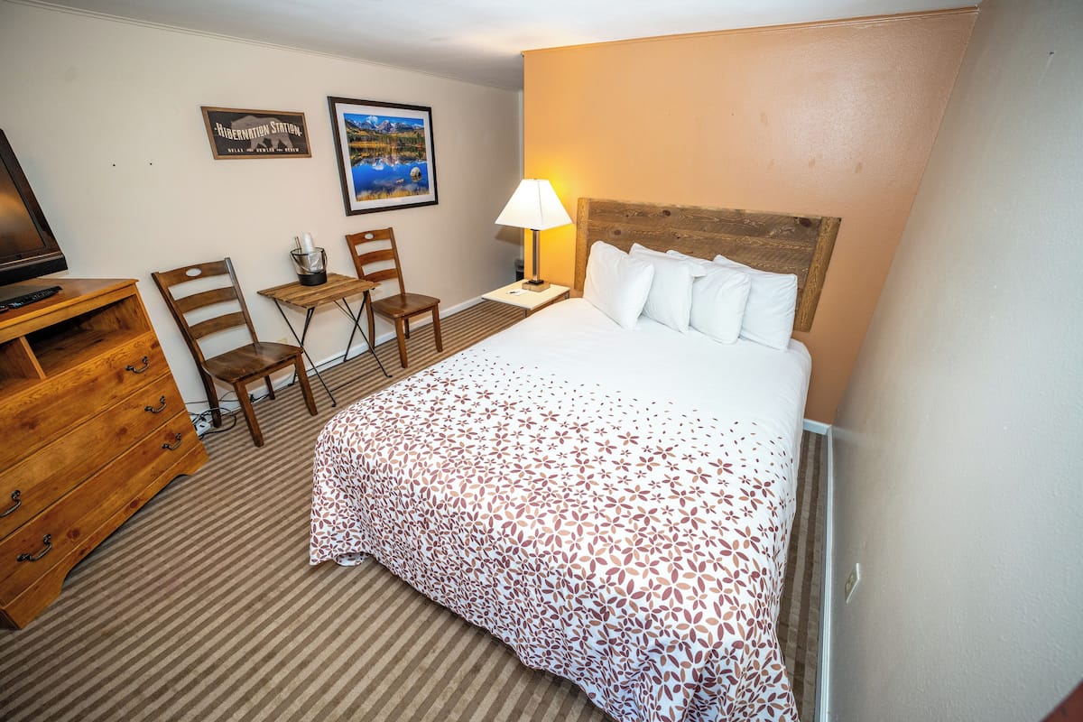 A hotel room with a bed, a desk, two chairs, a lamp, and a television.
