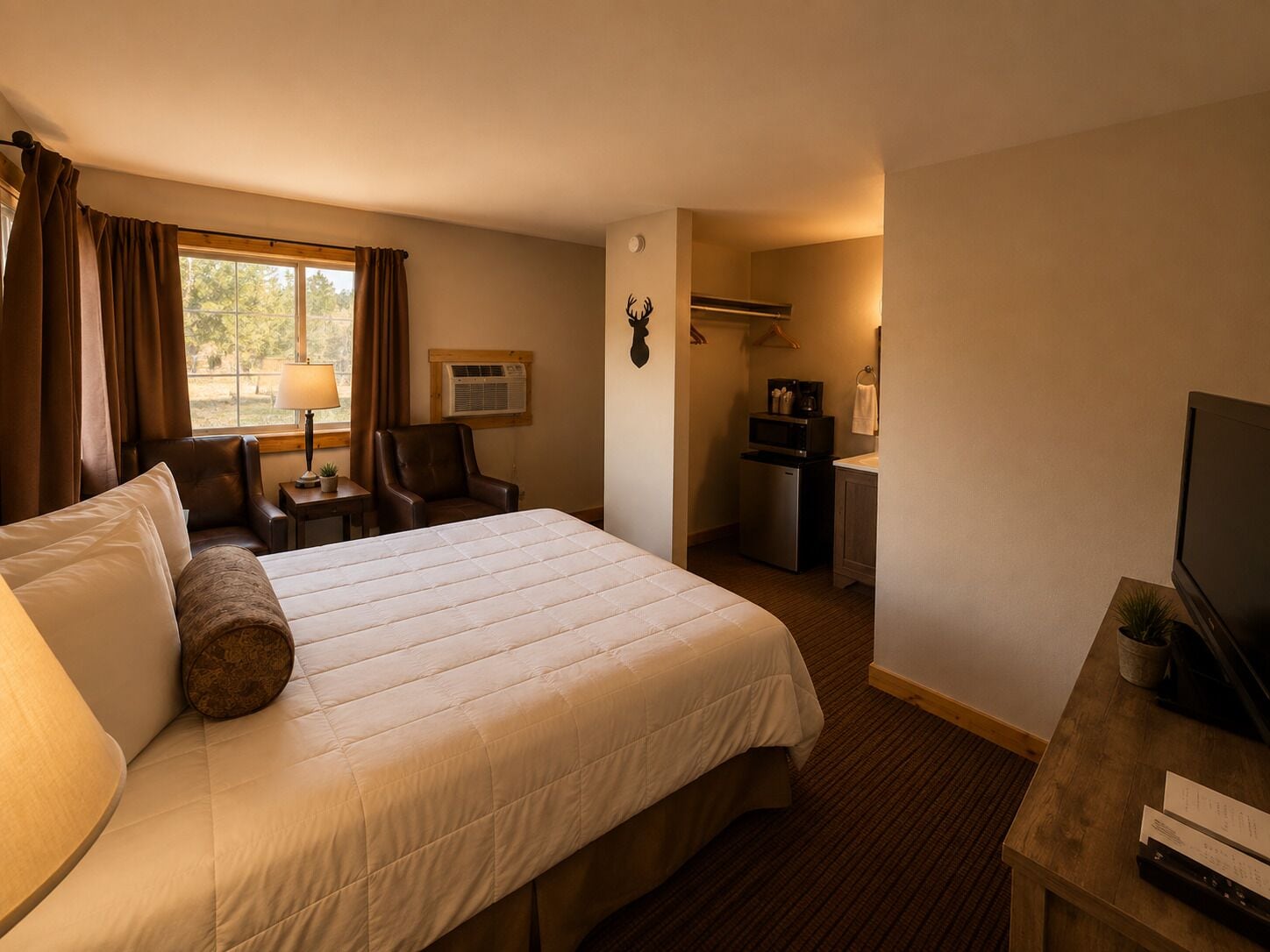A hotel room with a large bed, a desk with a television, a small kitchenette, and a window with curtains.