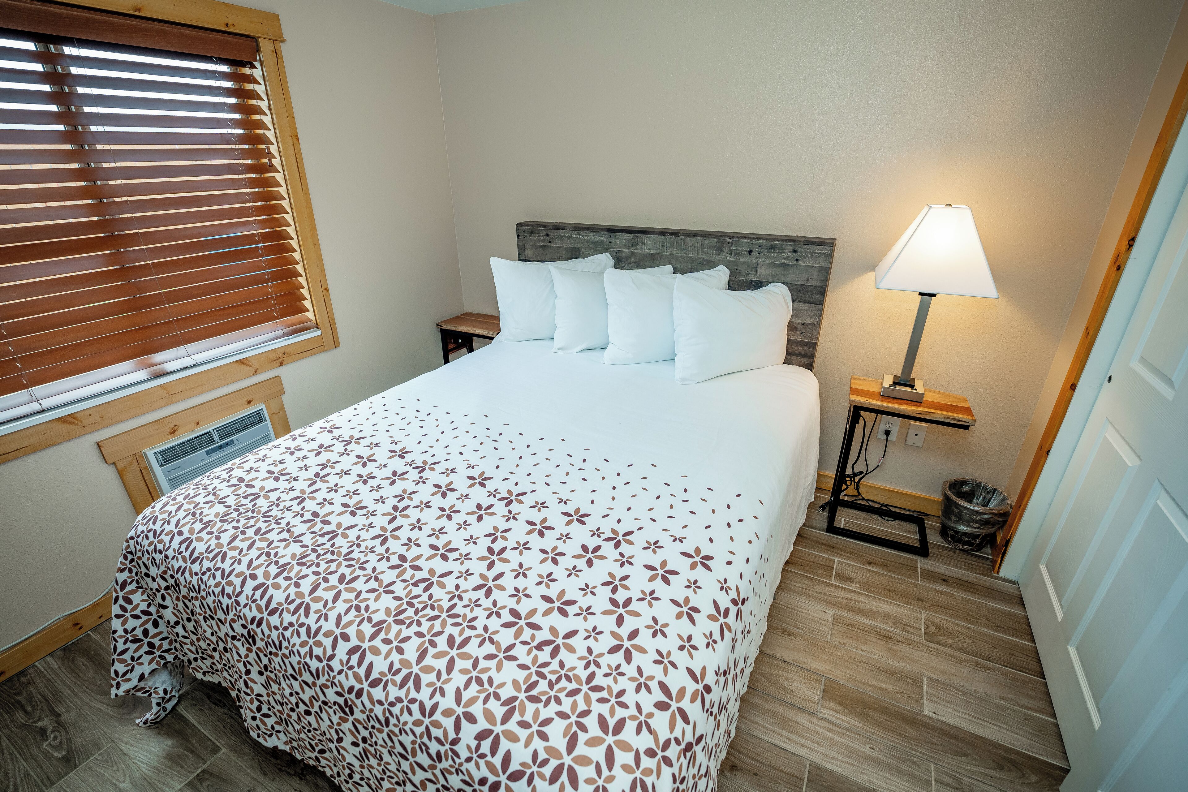 A neatly made bed with a patterned bedspread, a bedside lamp, and a window with blinds.