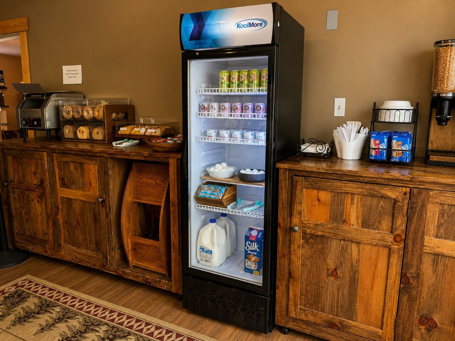 A self-service beverage dispenser with various drinks and a milk cooler, located in a hotel lobby with wooden furniture and a coffee machine.
