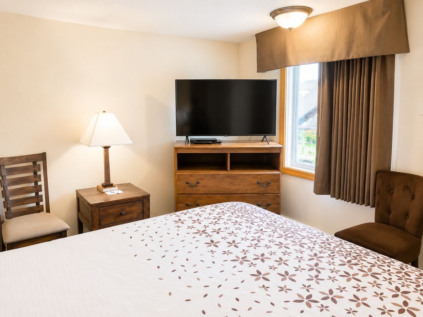 A hotel room with a bed, a TV on a wooden stand, a lamp, a chair, and a window with curtains.