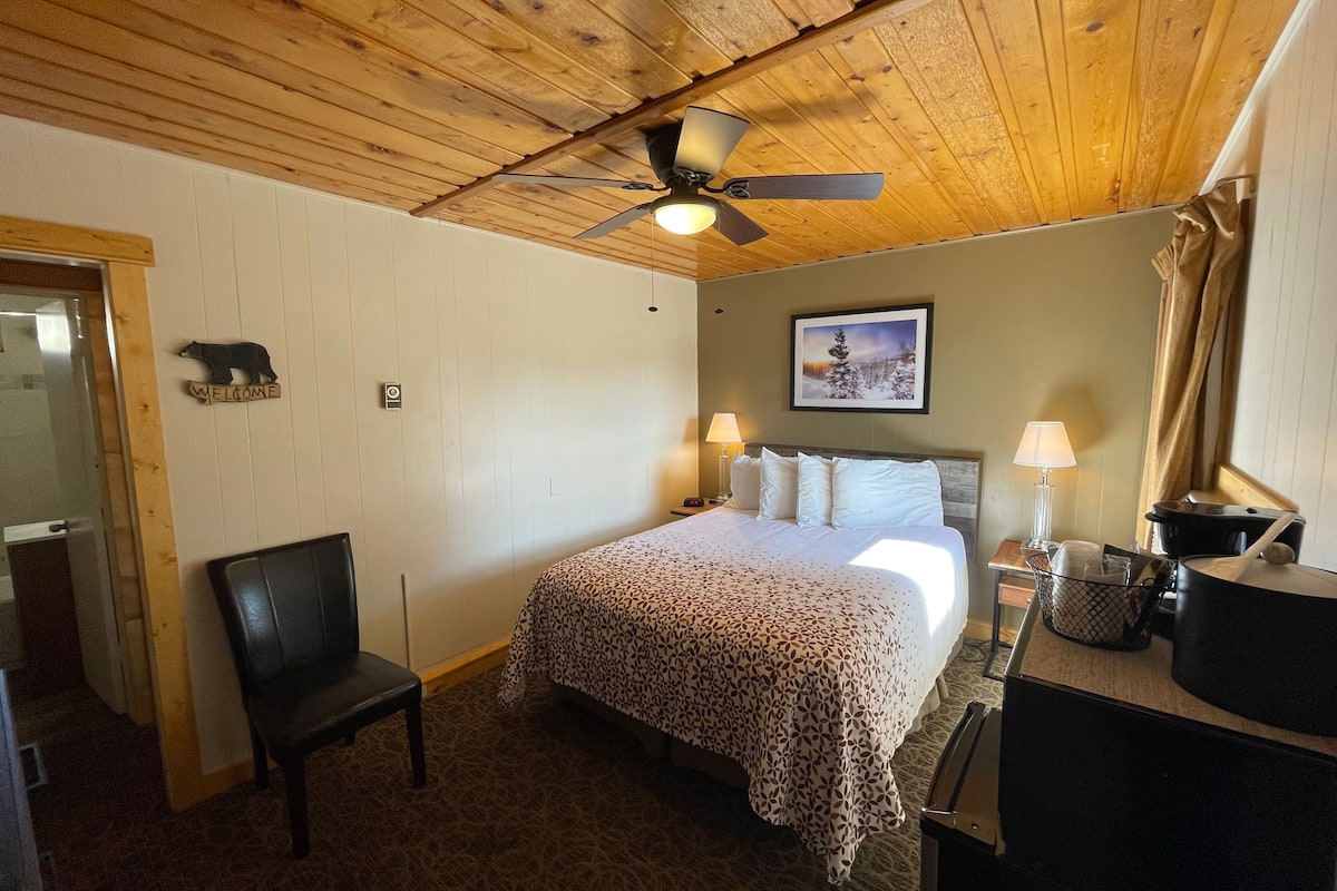 A bedroom with a wooden ceiling, a bed with a leopard print bedspread, two bedside lamps, a framed picture on the wall, and a black chair.
