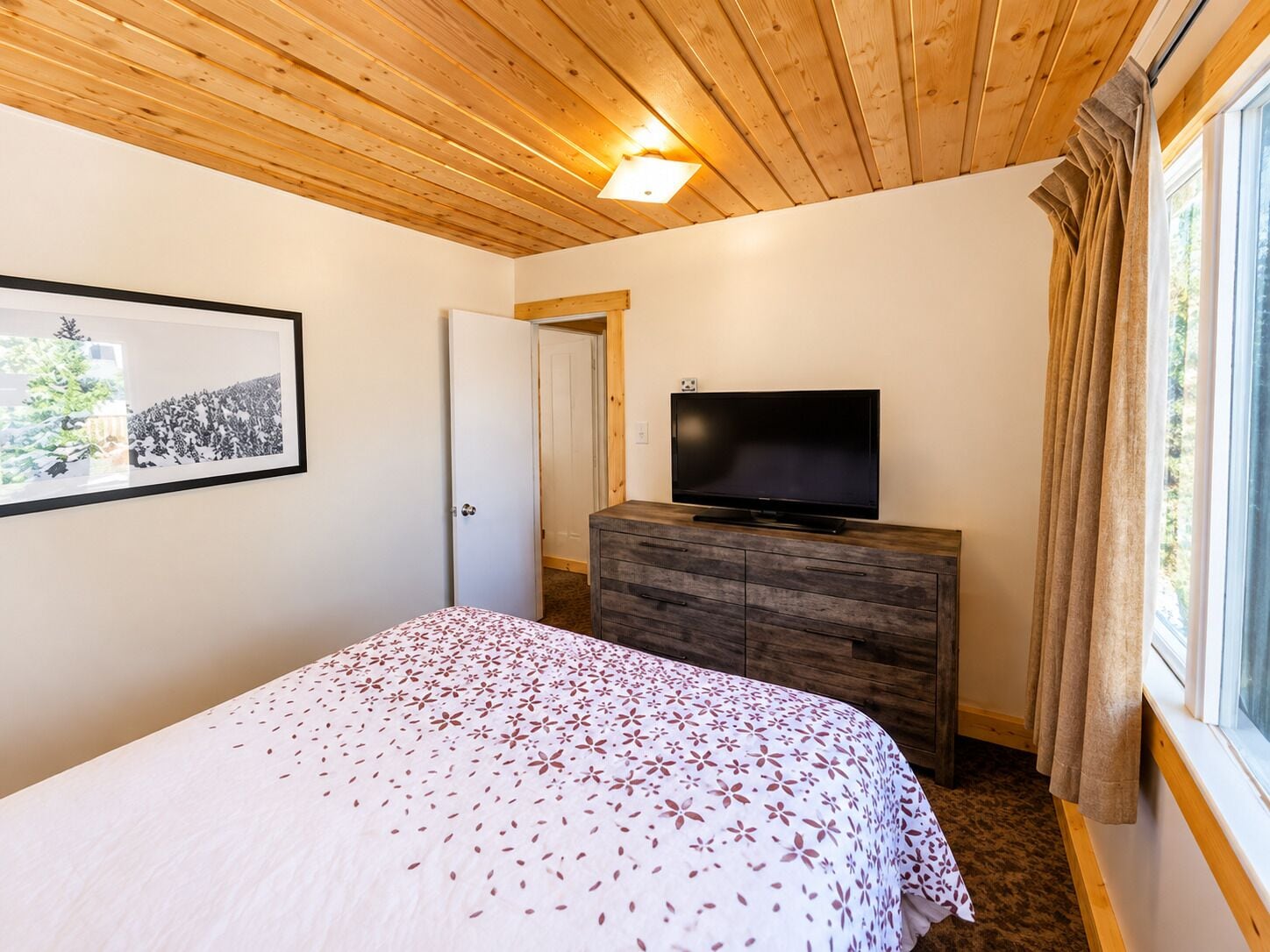 A bedroom with a bed, a television on a wooden dresser, a framed picture on the wall, and a window with curtains.