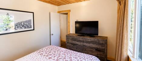 A bedroom with a bed, a television on a wooden dresser, a framed picture on the wall, and a window with curtains.