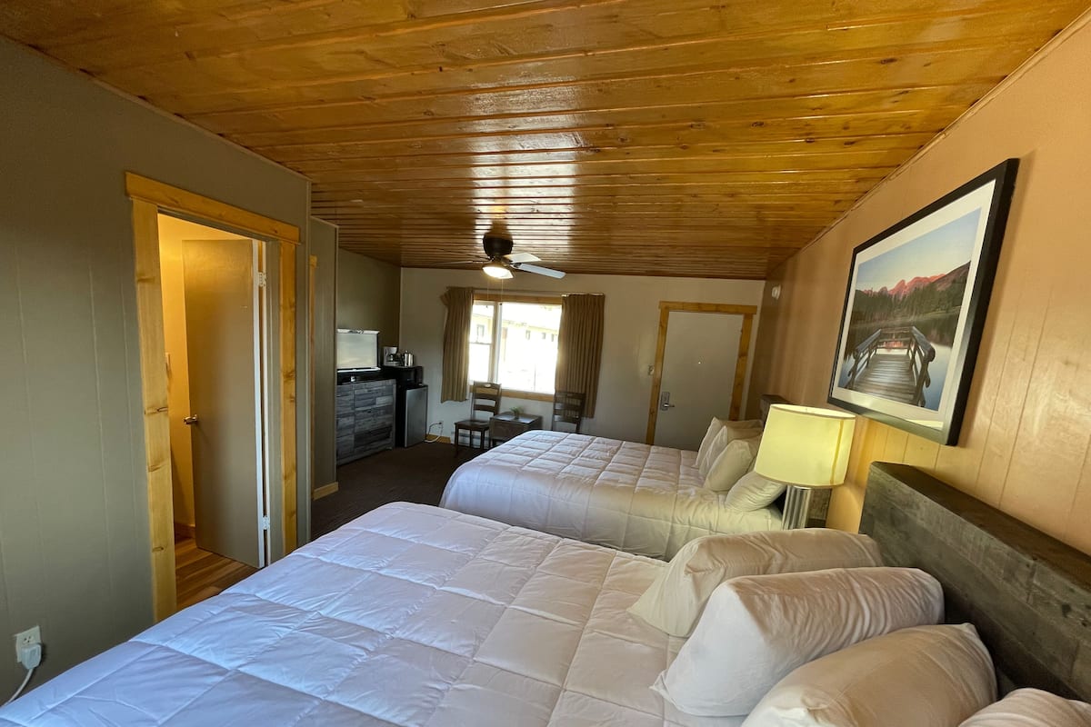 A hotel room with two beds, a desk, a chair, a small kitchenette, and a ceiling fan.