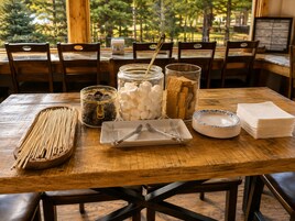 Una mesa de madera para comer con diferentes alimentos y utensilios, rodeada de sillas.