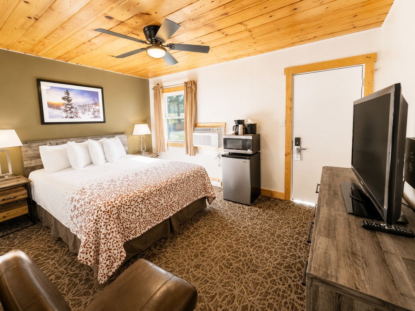 Habitación de hotel con cama, televisor, refrigerador, escritorio y ventilador de techo.