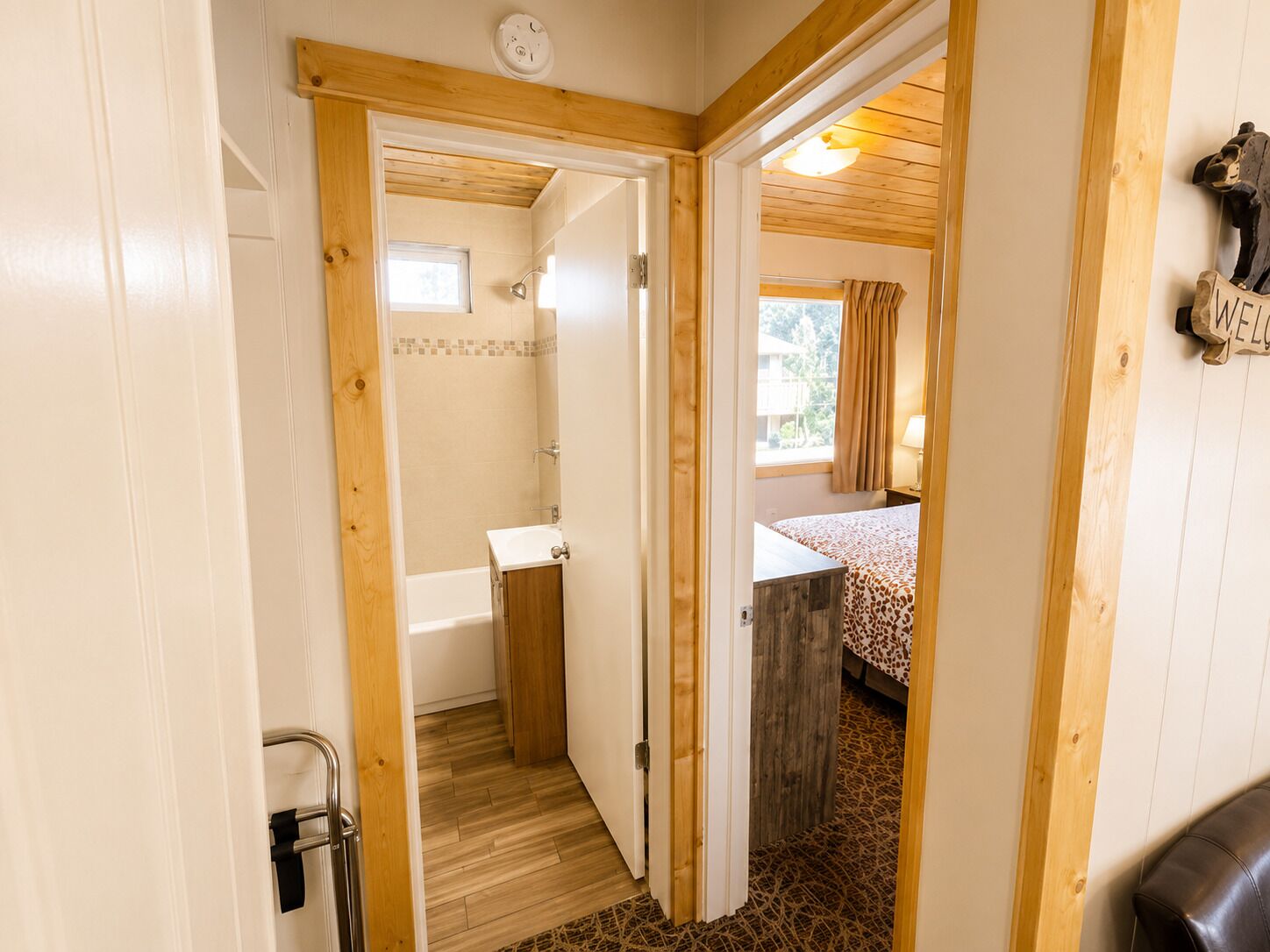 Ein Zimmer mit Holztür zu einem Badezimmer, einem Bett mit gemustertem Überwurf und einem Willkommensschild an der Wand.