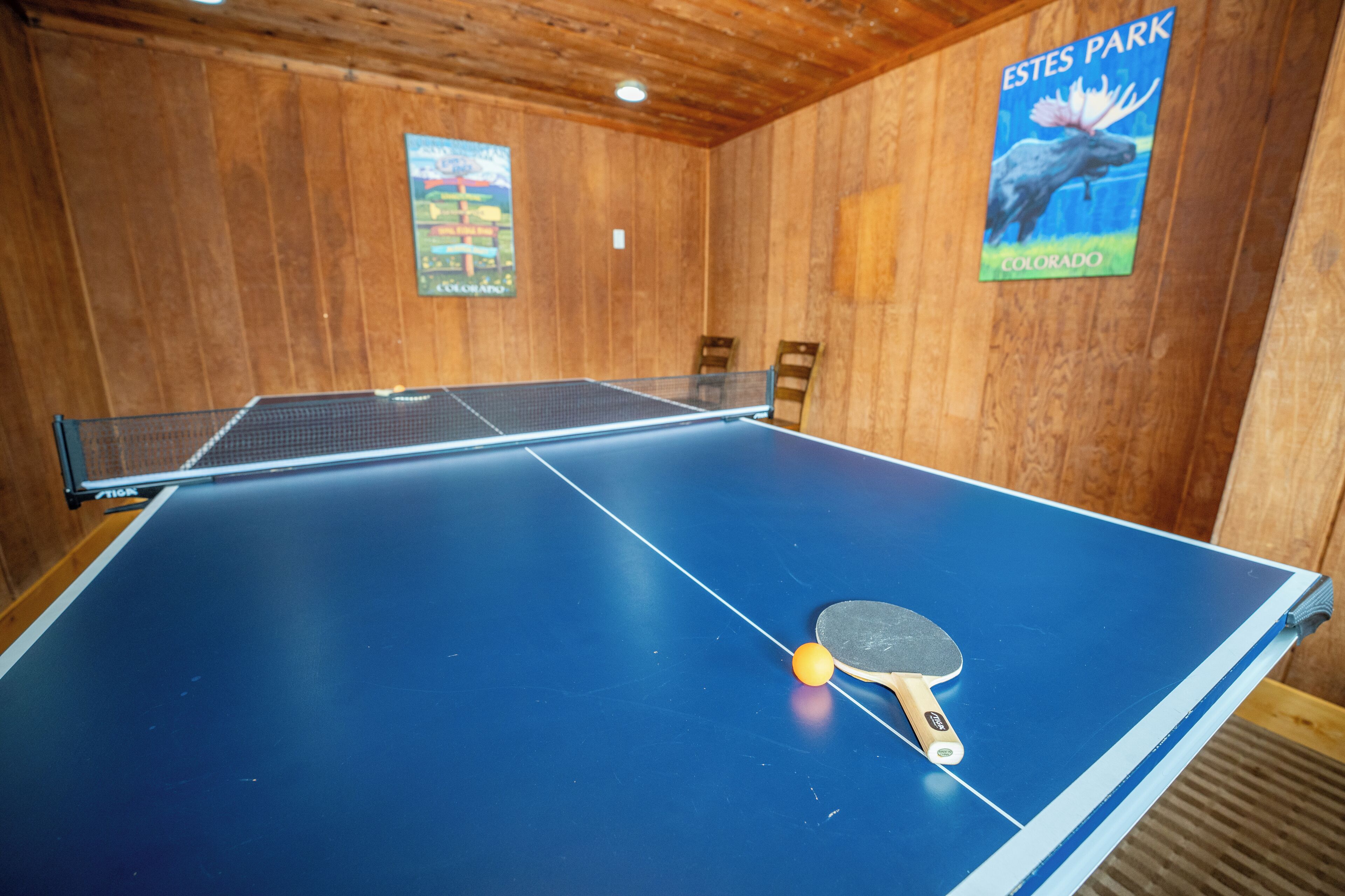 A ping pong table with a paddle and ball in a room with wooden walls and a poster of a moose.