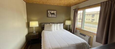 A hotel room with a wooden ceiling, a bed with white bedding, two bedside lamps, a window with curtains, and a picture of a deer on the wall.