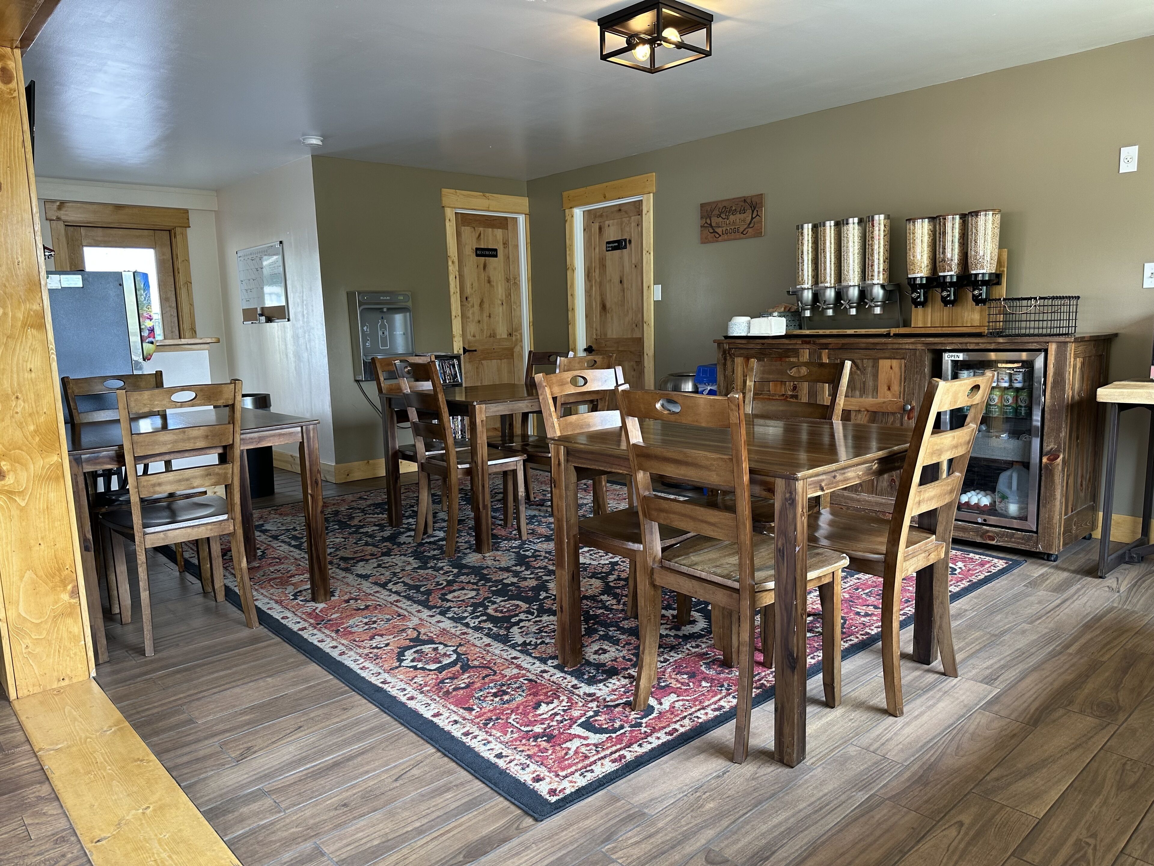 Un comedor con mesas y sillas de madera, un tapete estampado y un refrigerador con bebidas.