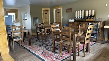 A dining area with wooden tables and chairs, a patterned rug, and a refrigerator with beverages.