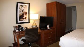 A hotel room with a bed, a desk with a chair, a television, a lamp, a framed picture on the wall, and a wardrobe.