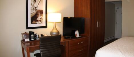 A hotel room with a bed, a desk with a chair, a television, a lamp, a framed picture on the wall, and a wardrobe.